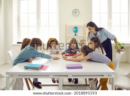 Happy school teacher and first grade students having interesting engaging activities in class. Little children sitting around big classroom table, learning ABCs, talking and playing fun games together