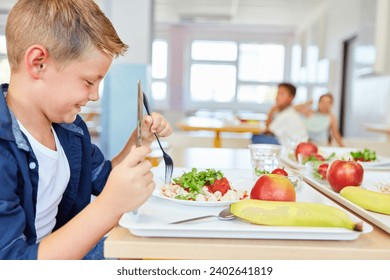 Happy schoolboy eating meal with cutlery while sitting at table in cafeteria - Powered by Shutterstock