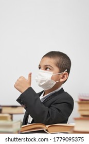 Happy Schoolboy In Face Mask. Mixed-race Kid Shows Thumbs-up Gesture Between Pile Of Books. Back To School After Covid-19. School Year Start After Long Delay Due To Coronavirus