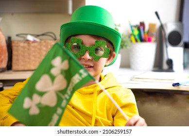 Happy Schoolboy On St. Patrick Day. Wearing A Green Hat, Wearing Green Clover-shaped Glasses, Waving A Small Green Flag. At Home