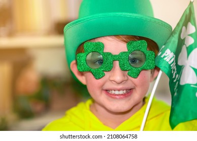 Happy Schoolboy On St. Patrick Day. Wearing A Green Hat, Wearing Green Clover-shaped Glasses, Waving A Small Green Flag. At Home