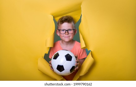 Happy schoolboy with soccer ball. and breaking through yellow paper wall. Happy smiling kid go back to school, kindergarten. Success, motivation, winner concept. Little boy dreaming to be super hero.
