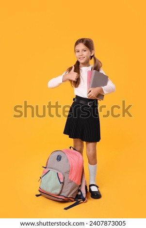Happy schoolgirl with backpack and books showing thumb up on orange background