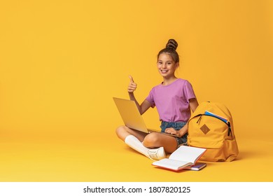 happy schoolgirl with backpack, laptop and books is sitting on yellow background in the Studio. The child shows, a sign of approval, a finger up. Back to school. copy space.