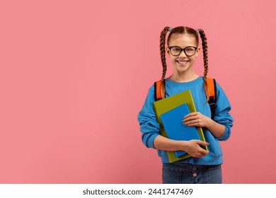 A happy schoolgirl with braids holding blue books and wearing a backpack smiles at the camera - Powered by Shutterstock