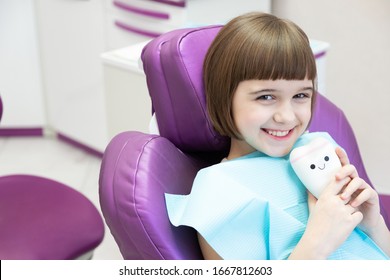 Happy  Schoolgirl  In Dental Chair. Cute Small Girl Holding Toy Tooth In Hand. Dental Office In Modern Pediatric Clinic. Teeth Treatment For Children. Kid Medical Insurance