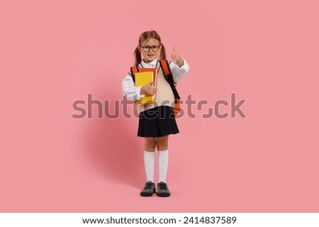 Happy schoolgirl in glasses with backpack and books showing thumb up gesture on pink background