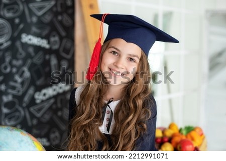 Happy Schoolgirl in master's hat. Portrait of happy little girl in graduate headdress. Smiling Little girl in a school uniform in classroom at school. Back to school. A joyful beautiful graduate. 
