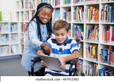 Happy schoolgirl standing with schoolboy on wheelchair using digital tablet in library - Powered by Shutterstock