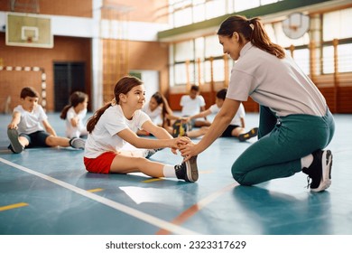 Happy schoolgirl stretching her leg with teacher's assistance on physical education class at school gym.