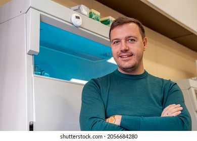 Happy Scientist Poses In Room With Laboratory Box In UV Light For Sterilization In Background. Young Successful Male Worker Of Scientific Laboratory In Casual Wear Sitting By Laboratory Shelf