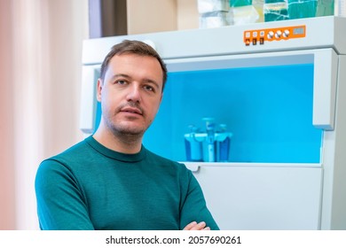 Happy Scientist Poses In Room With Laboratory Box In UV Light For Sterilization In Background. Young Successful Male Worker Of Scientific Laboratory In Casual Wear Sitting By Laboratory Shelf Inside