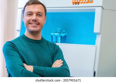 Happy Scientist Poses In Room With Laboratory Box In UV Light For Sterilization In Background. Successful Male Worker Of Scientific Laboratory Sitting By Laboratory Shelf Inside Office In Front Of Cam