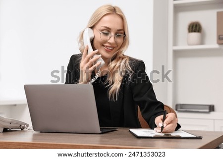 Happy secretary taking notes while talking on phone at table in office