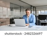 Happy senior 50 years old professional business man wearing suit using computer sitting at desk. Busy smiling middle aged businessman executive investor bank manager working on computer in office.