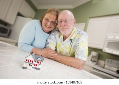 Happy Senior Adult Couple Gazing Over Small Model Home On Their Kitchen Counter.