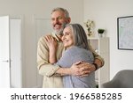 Happy senior adult mature classy couple hugging, bonding, thinking of good future. Carefree cheerful mid age old husband embracing wife looking away dreaming, enjoying wellbeing and love in new house.