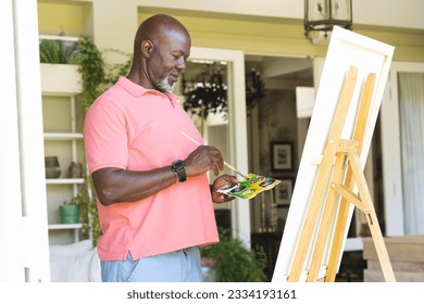 Happy senior african american man holding paint palette painting canvas on easel at home. Senior lifestyle, retirement, creativity, hobbies, art and domestic life, unaltered. - Powered by Shutterstock