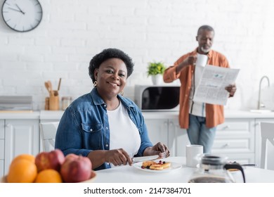 Happy And Senior African American Woman Having Breakfast Near Blurred Husband With Newspaper