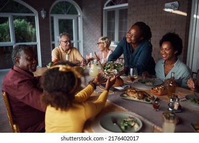 Happy Senior African American Woman Serving Food To Her Multiracial Family At Dining Table On A Patio. 