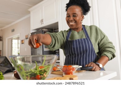 Happy senior african american woman making salad and using tablet in kitchen at home. Domestic life, retirement, food, technology and lifestyle, unaltered. - Powered by Shutterstock