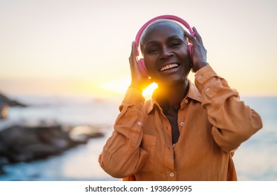 Happy senior African woman enjoying listening music in front of sea during summer sunset