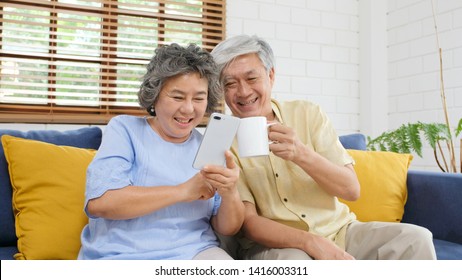 Happy Senior Asian Couple Using Smart Phone While Sitting On Sofa At Home Living Room Background, Active Senior People Retirement And Technology Lifestyles