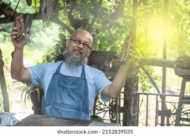 Happy Senior Asian Leather-making Man In Workshop Clothes Is Holding Leather Products He Made, An Aged Artistic Handicraftsman Shows Handmade Leather Crafting Bags On Hands, Art And Craft Concept