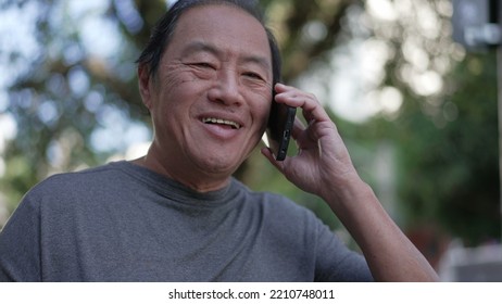 Happy Senior Asian Man Speaking On Phone While Walking In City Street. Portrait Of An Older Middle Aged Person Talking In Conversation With Cellphone Device