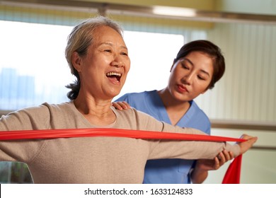 Happy Senior Asian Woman Exercising Using Resistance Band Guided By Physical Therapist In Rehab Center