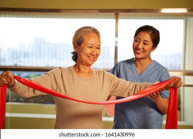 Happy Senior Asian Woman Exercising Using Resistance Band Guided By Physical Therapist In Rehab Center