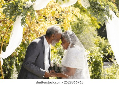 Happy senior biracial bride and groom during wedding ceremony in sunny garden. Marriage, love, romance, ceremony, summer, tradition and senior lifestyle, unaltered. - Powered by Shutterstock