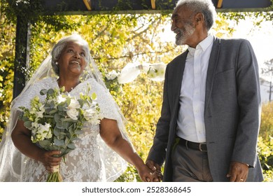 Happy senior biracial bride and groom holding hands at sunny outdoor wedding ceremony. Marriage, romance, love, tradition, ceremony, summer, retirement and senior lifestyle, unaltered. - Powered by Shutterstock