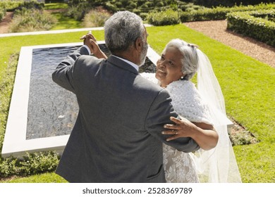 Happy senior biracial bride and groom dancing in garden at sunny wedding ceremony, copy space. Marriage, romance, love, tradition, ceremony, summer, retirement and senior lifestyle, unaltered. - Powered by Shutterstock