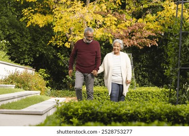 Happy senior biracial couple holding hands and walking in garden at home. Senior lifestyle, retirement, nature and domestic life, unaltered. - Powered by Shutterstock