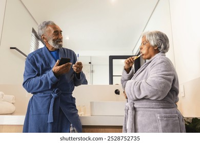 Happy senior biracial couple using smartphone and brushing teeth in bathroom at home. Senior lifestyle, retirement, technology, selfcare and domestic life, unaltered. - Powered by Shutterstock