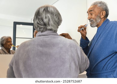 Happy senior biracial couple wearing bathrobes and brushing teeth in bathroom at home. Senior lifestyle, retirement, selfcare and domestic life, unaltered. - Powered by Shutterstock