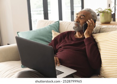 Happy senior biracial man sitting on couch with laptop talking on smartphone at home. Senior lifestyle, retirement, technology, communication and domestic life, unaltered. - Powered by Shutterstock