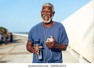 A happy senior Black man runs along a seaside promenade holding a water bottle and smartphone, wearing earphones and a joyful expression. - Powered by Shutterstock