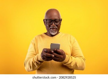 Happy senior black man using smartphone, writing message, chatting with friend, surfing internet, watching movie online, having webinar or meeting on orange studio background