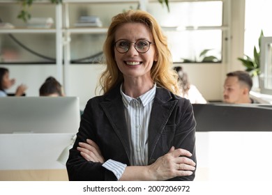 Happy senior business woman, leader, manager in glasses looking at camera with hands folded, ream working behind. Female mentor, coach, teacher posing in office with group. Head shot portrait