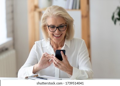 Happy senior businesswoman in glasses sit in office text message on smartphone gadget, smiling middle-aged female employee browse wireless Internet on cell device at workplace, technology concept