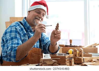Happy senior caucasian man wearing Santa hat holds and paints small wooden Christmas tree, prepare toys ready for Christmas