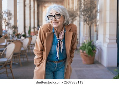Happy senior Caucasian woman poses confidently in an outdoor cafe space. Dressed in a brown coat, scarf, and glasses, she expresses elegance, experience, and timeless fashion in daylight. 