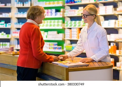 Happy Senior Citizen Customer In Red Standing At Pharmacy Counter As Pharmacist In Eyeglasses And Lab Coat Hands Her A Medication Order