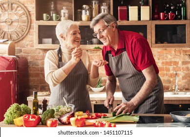 Happy Senior Couple In Aprons Tasting Food And Smiling, Cooking Healthy Lunch Together In Kitchen