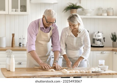 Happy senior couple of bakers enjoying culinary hobby in home kitchen, rolling raw dough, talking, smiling, laughing, preparing dessert for family dinner, baking fresh buns - Powered by Shutterstock