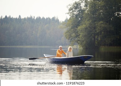 Happy senior couple in casual clothing sitting on boat and enjoying romantic date on lake, elderly man rowing oars