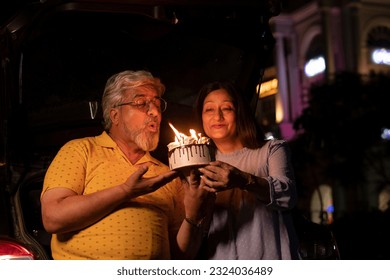 Happy senior couple celebrating birthday and cutting cake at night standing with car at public place - Powered by Shutterstock