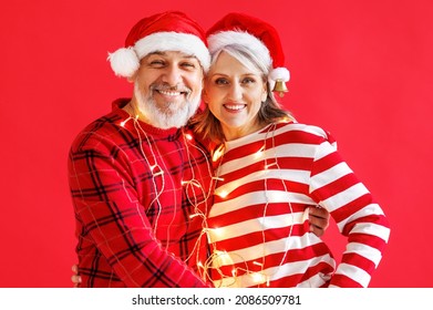 Happy senior couple in Christmas twinkling lights embrace on red background, smiling aged man and woman in Santa hats celebrate New Years eve together. Retired pensioners enjoy xmas holidays - Powered by Shutterstock